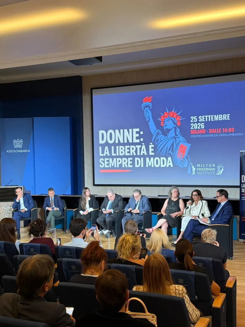 Platea piena e relatori sul palco sotto lo schermo con il titolo dell’evento
